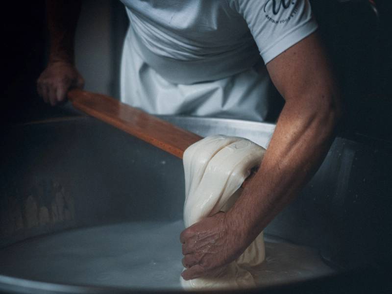 En person som strekker ut og drar i en stor klump med ost på et bord, sannsynligvis i et ysteri.