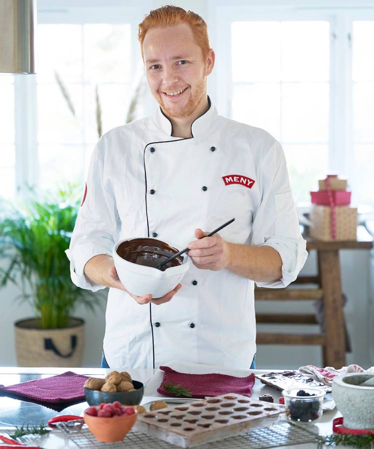 En person i kokkeforkle og hvit kokkeuniform holder en bolle med julekonfekt, med julepynt og bakverk på bordet foran seg.
