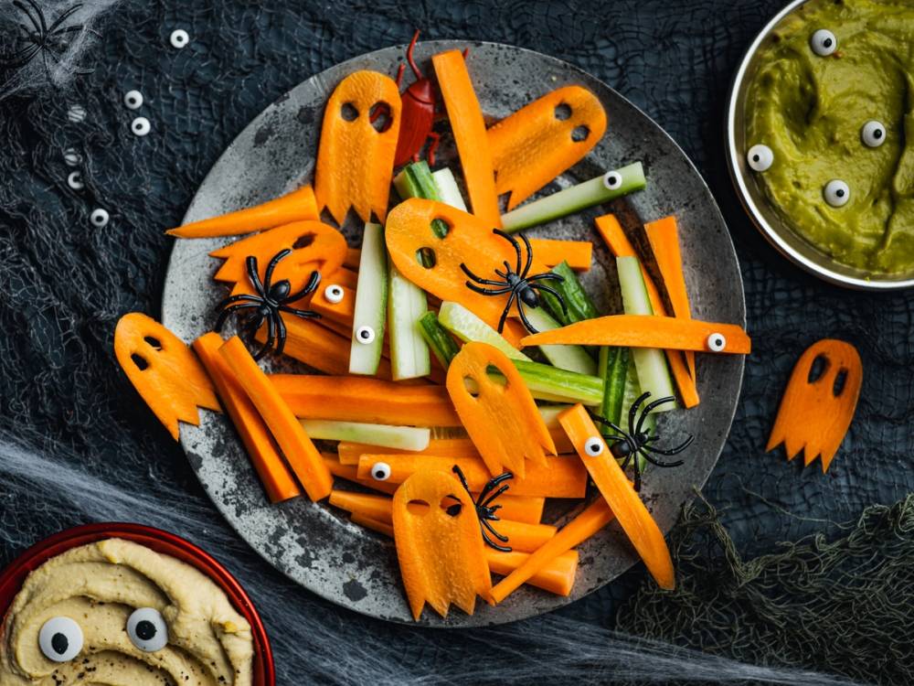 Skål med utskårne grønnsaker og dekorative spøkelser til Halloween, omgitt av små plastøyne og en skål med grønnsaker i olje