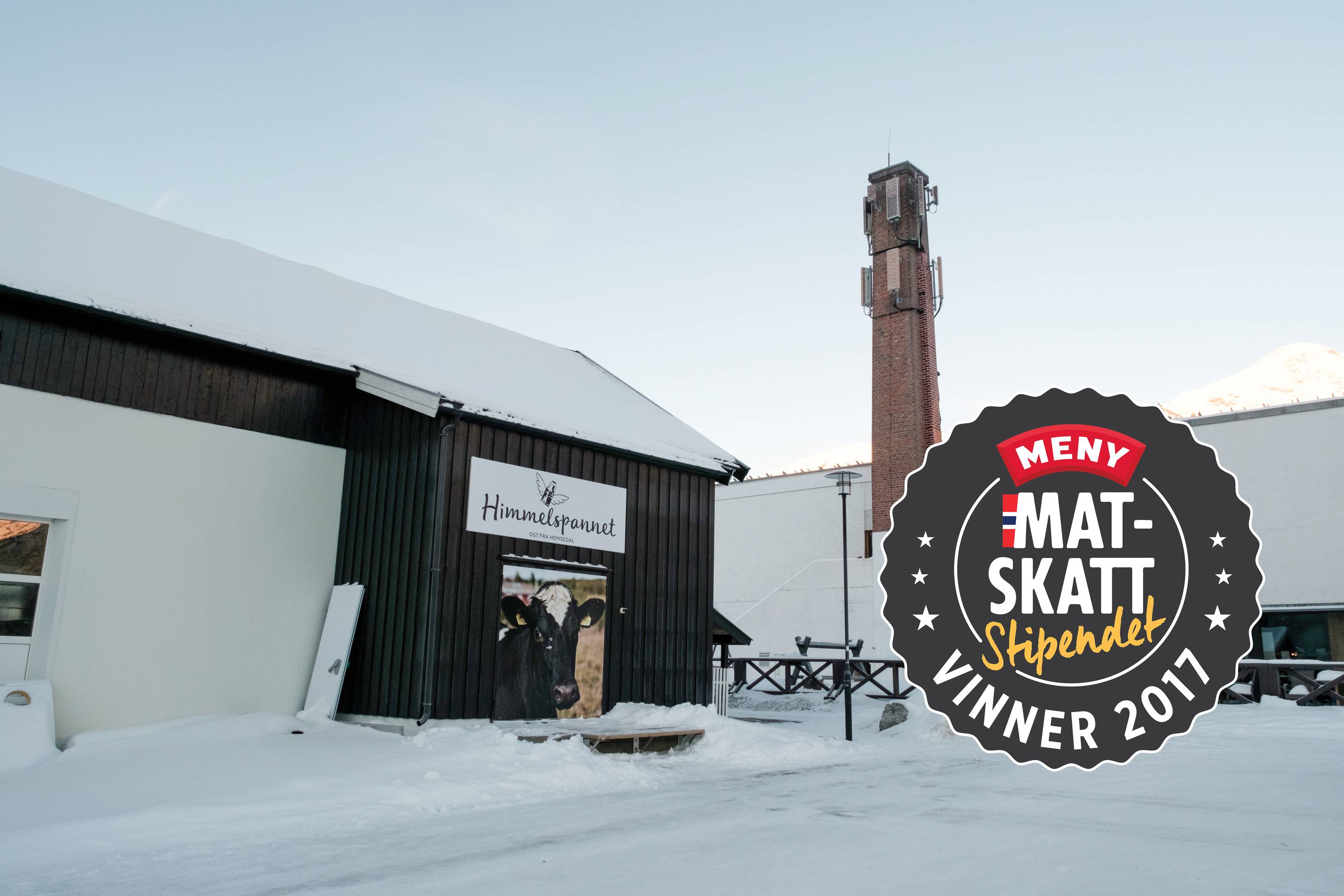 En bygning med skilt som sier 'Hjemme hos oss', omgitt av snø og en tårn i bakgrunnen, med en logo for 'Meny Mat Skatt' i hjørnet.