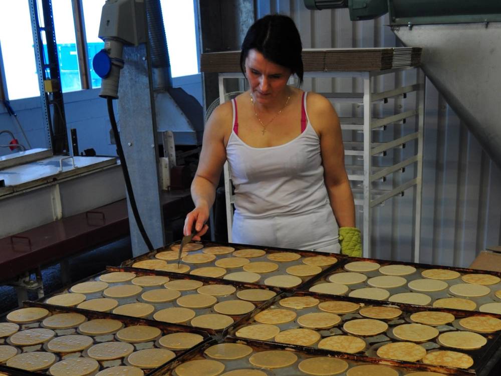 En person med svart hår og hvit topp står ved et bord med flere runde kaker eller kjeks, i et kjøkken eller bakeri.