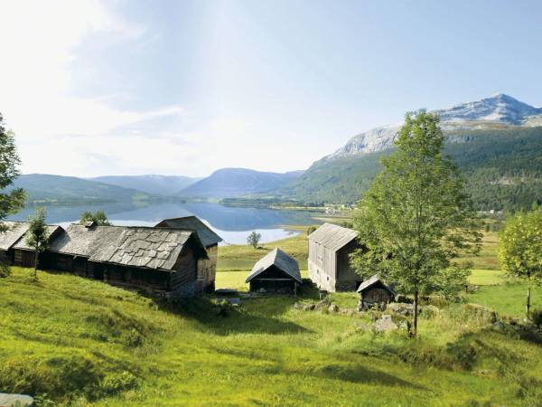 Utsikt over en norsk landsby med gamle trebygninger, grønne åser og fjell i bakgrunnen, med vann i forgrunnen.