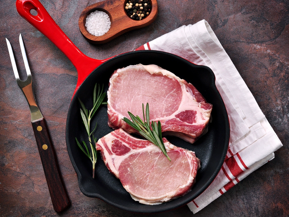 To rå koteletter med ben ligger i en svart panne, pyntet med en kvist rosmarin, på et mørkt bord med en hvit og rødstripet kjøkkenklut ved siden av.
