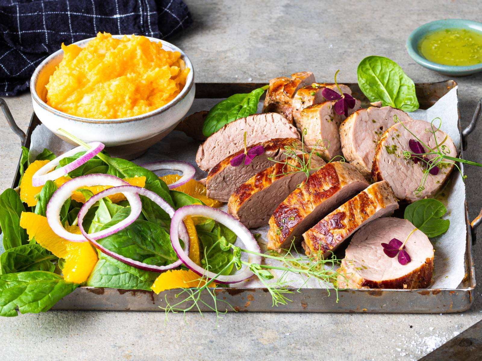 Svinefilet skåret i skiver med grønnsaker og potetmos på en tallerken, med en liten skål med grønn saus ved siden av.