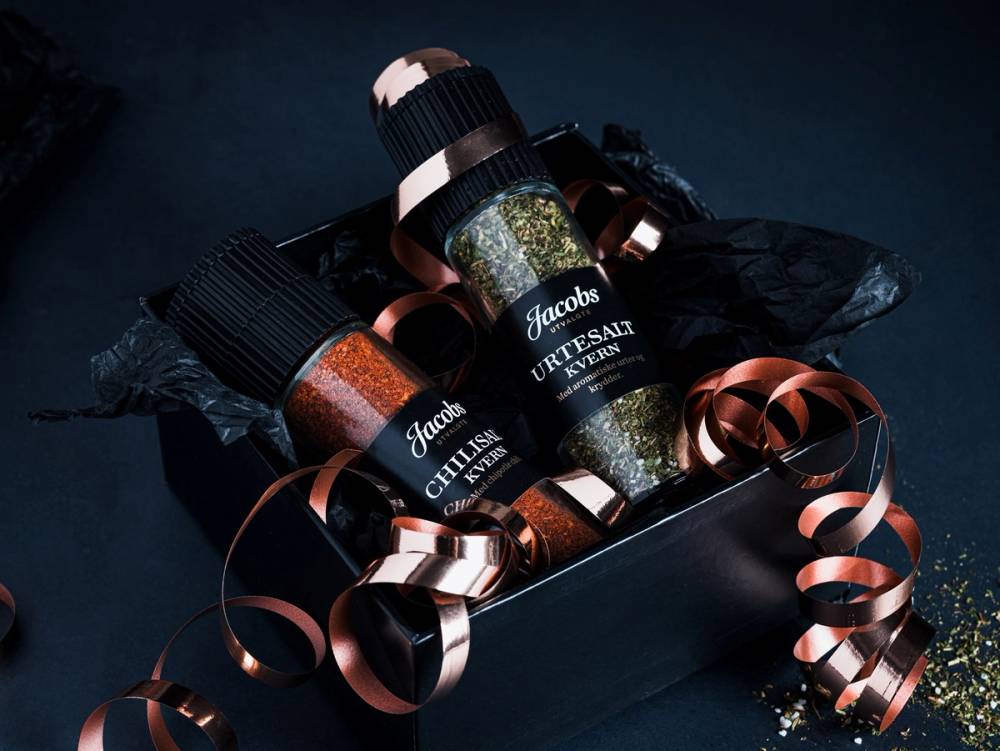 To små krydderglass med svart etikett og gullfargede detaljer, plassert i en svart eske med gullfarget bånd og konfetti rundt.