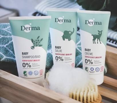 Tre babypleieprodukter fra Derma, inkludert sjampo, salve og krem, plassert i en kurv med en børste og en klut ved siden av.