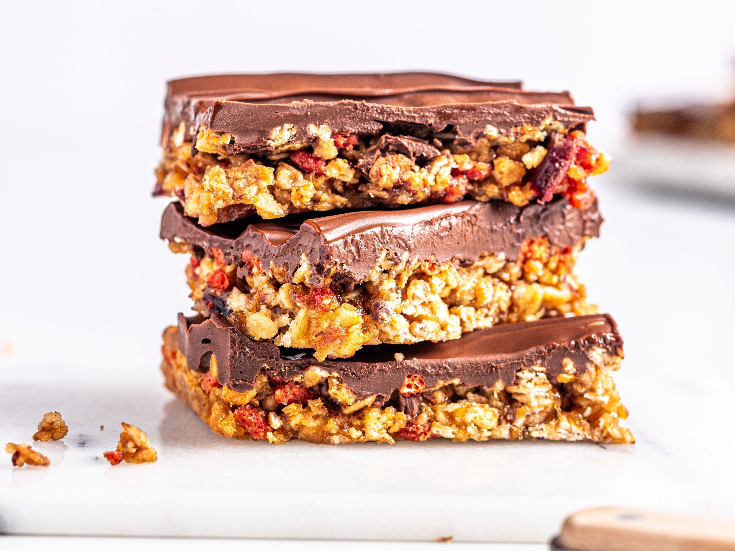 Tre sjokoladebelagte granola barer med nøtter og frukt, stablet oppå hverandre på et hvitt underlag.