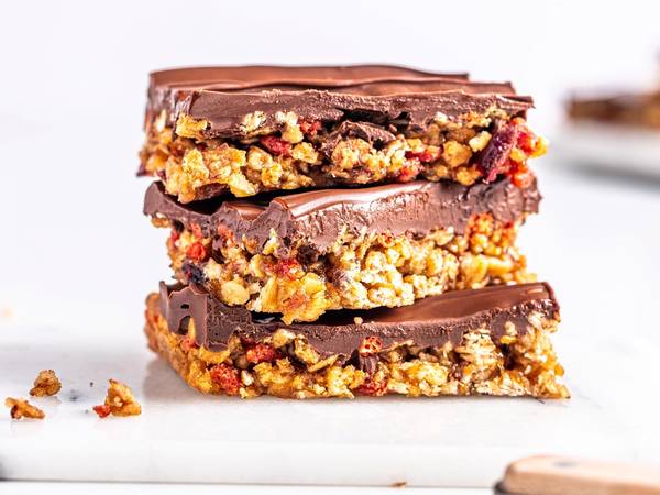 Tre sjokoladebelagte granola barer med nøtter og frukt, stablet oppå hverandre på et hvitt underlag.