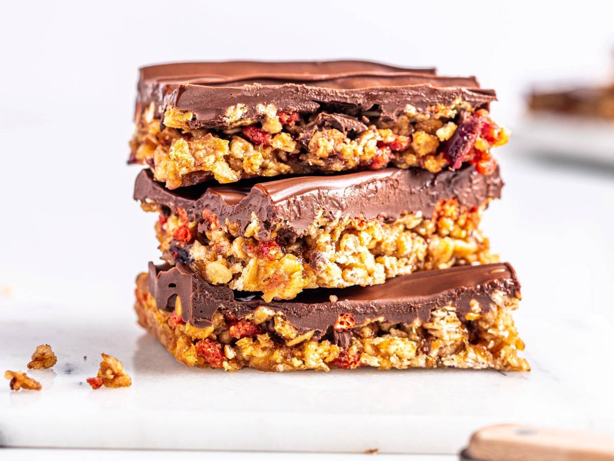 Tre sjokoladebelagte granola barer med nøtter og frukt, stablet oppå hverandre på et hvitt underlag.