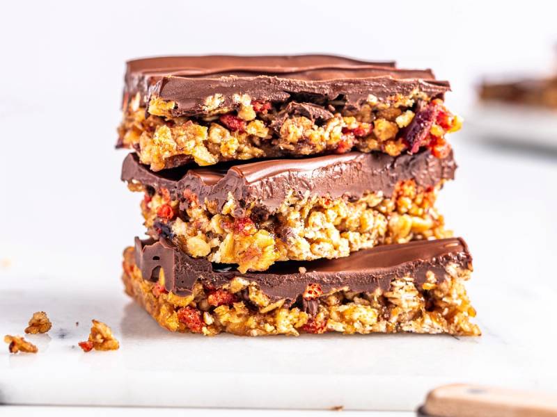 Tre sjokoladebelagte granola barer med nøtter og frukt, stablet oppå hverandre på et hvitt underlag.