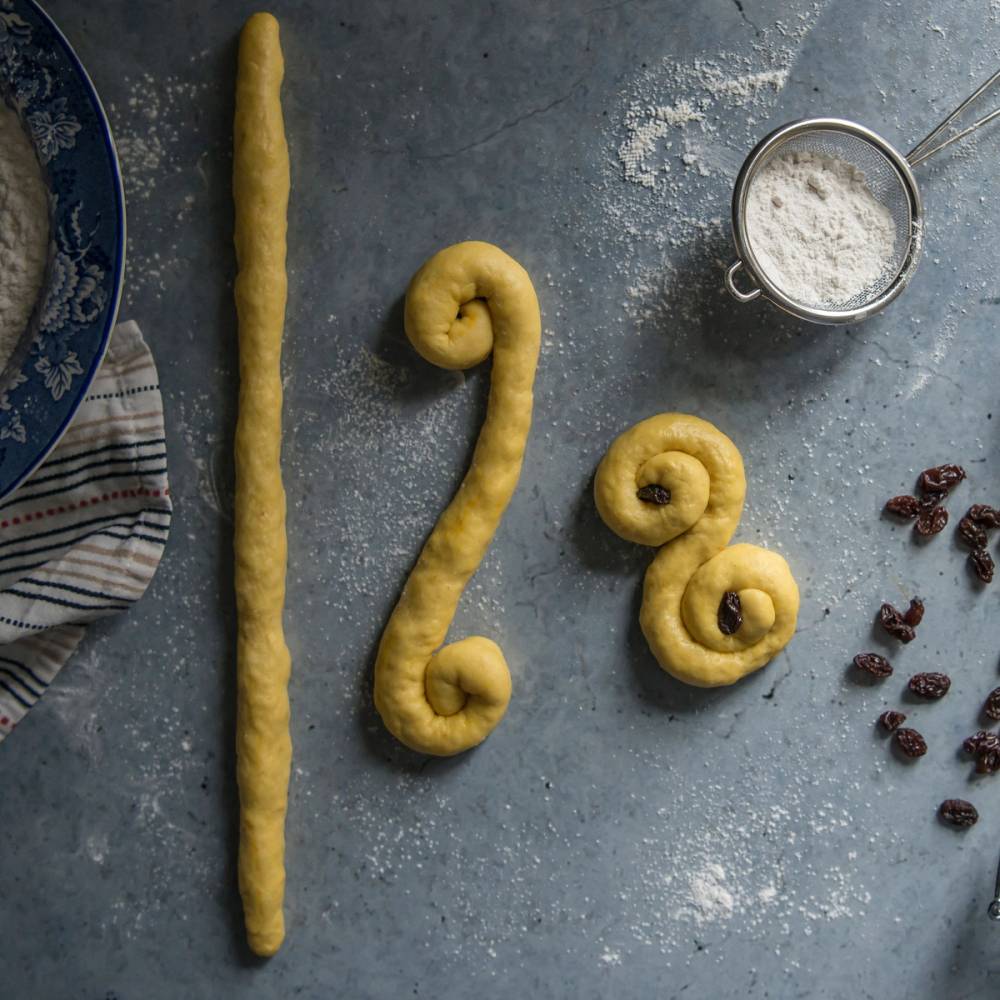 Lussekatt og julegris laget av deig, plassert på en kvadratisk overflate med mel og rosiner rundt.
