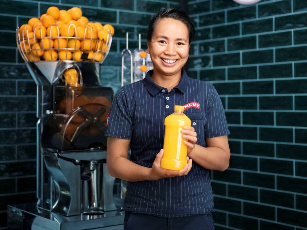 MENY-ansatt holder en flaske nypresset appelsinjuice foran juicepressen i butikk.