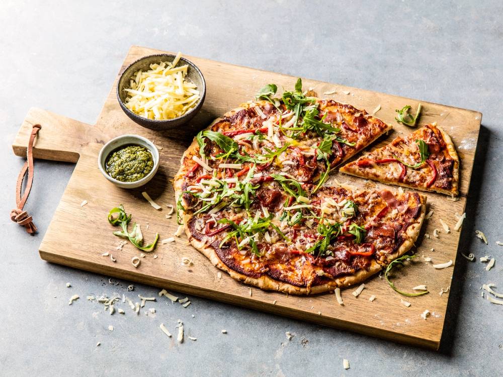 En pizza med ost, kjøtt og grønnsaker på en skjærebrett med en liten skål med pesto og en annen med revet ost ved siden av.