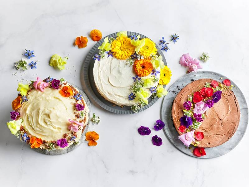 Tre kaker pyntet med ulike fargerike blomster på hvit bakgrunn.