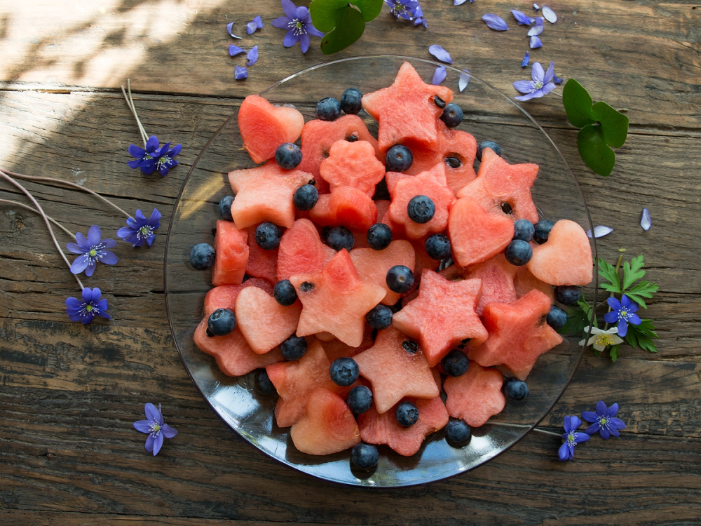 Fargerike vannmelonformede fruktbiter med blåbær på en glassplate, omgitt av blå blomster på et treverk