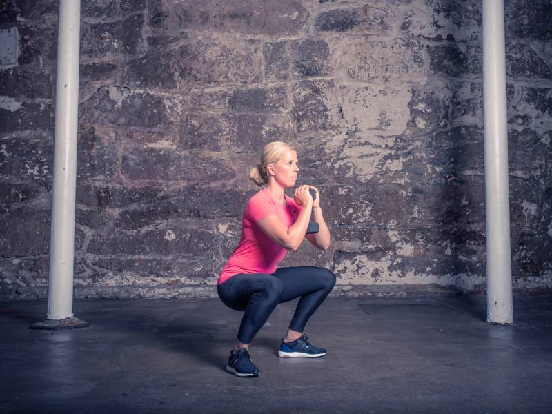 Kvinne i rosa treningstøy gjør en front knebøy i et treningsrom med murvegg og stenger på hver side.