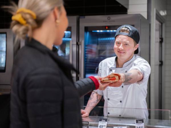 Kunde mottar mat fra ansatt i en butikk eller restaurant, med kjøleskap i bakgrunnen.