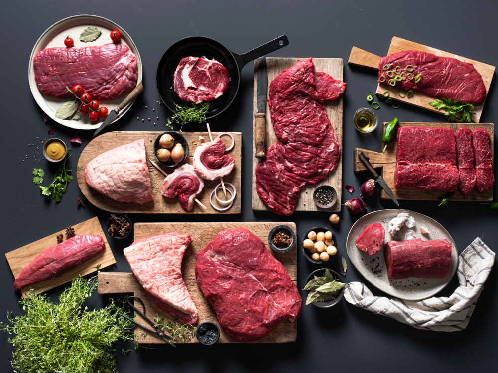 Ulike utvalg av rått kjøtt, inkludert biffer og steker, på forskjellige skjærebrett og tallerkener med urter og krydder rundt.
