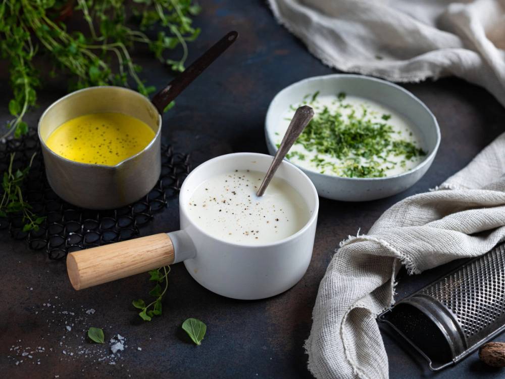 Tre små boller med hvit saus og en skje i hver, plassert på en mørk overflate med grønne urter og en hvit klut i nærheten.