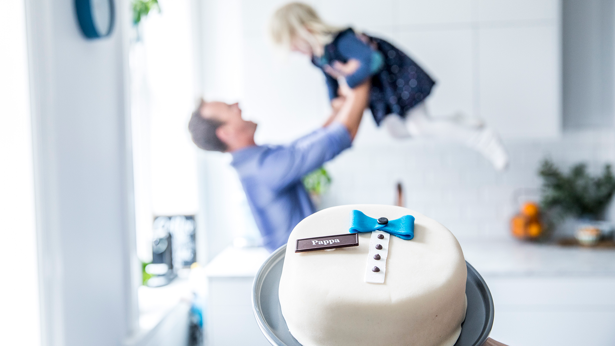 En far holder opp sin lille datter i luften over en bursdagskake i et kjøkken, feirer farsdag.