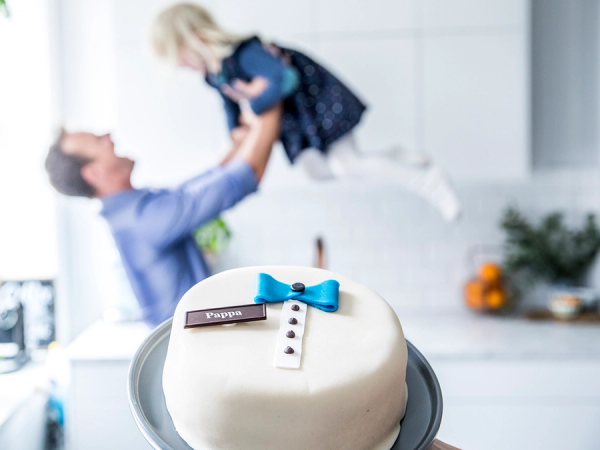 En far holder opp sin lille datter i luften over en bursdagskake i et kjøkken, feirer farsdag.