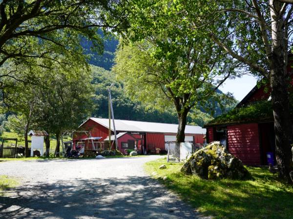 En gårdsplass med trær, en rød låve og en liten bygning, omgitt av grønt landskap og fjell i bakgrunnen.