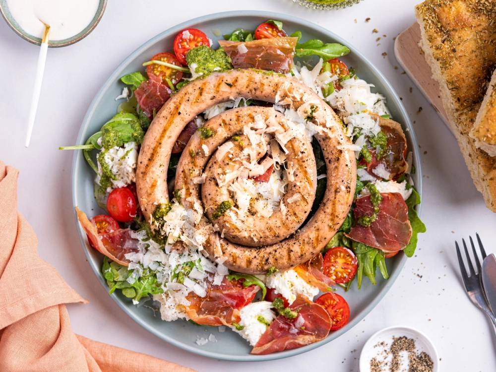 Salat med salsiccia, mozzarella, parmaskinke, cherrytomater og grønt på en hvit tallerken.