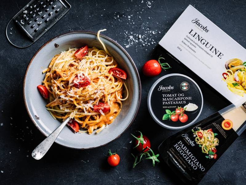 En tallerken med pasta med tomat og mozzarella, omgitt av cherrytomater og pastasaus, med pastapakker og en rivjern i bakgrunnen.