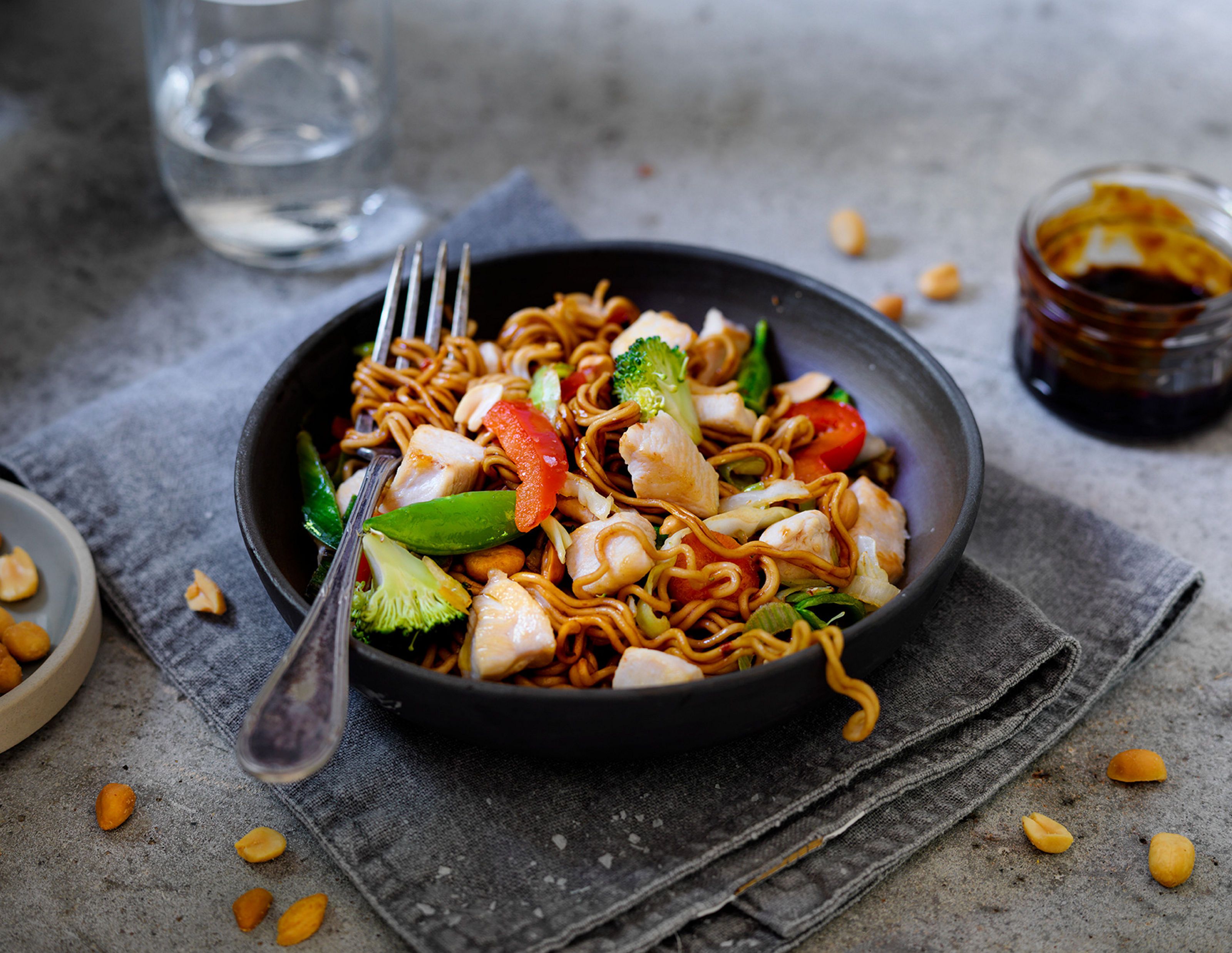 En tallerken med spicy kyllingwok med grønnsaker og nudler, med en skål med saus og et glass vann i bakgrunnen.