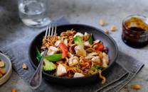 En tallerken med spicy kyllingwok med grønnsaker og nudler, med en skål med saus og et glass vann i bakgrunnen.