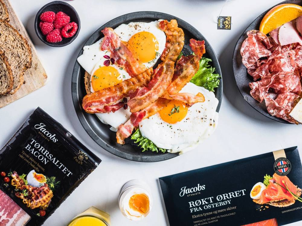 Frokost med stekte egg, bacon og grønnsaker på en tallerken, omgitt av brød, kjøtt og påskeegg, på et bord med ulike matvarer og pakker.