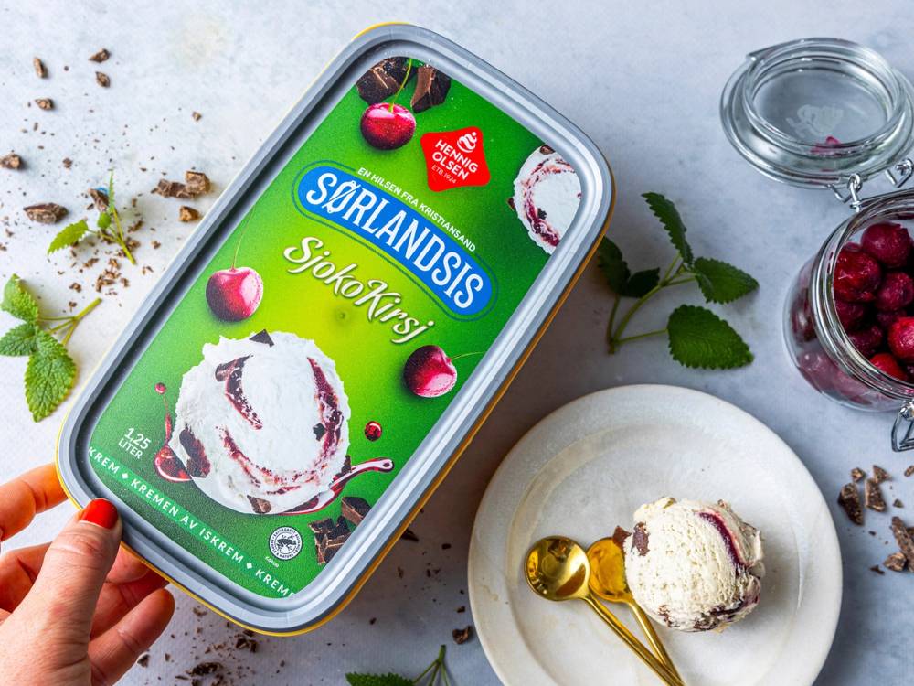 HennigOlsen sjokokirs is på en mobilskjerm med bilde av iskrem med sjokoladesmak og kirsebær, omgitt av kirsebær og mintblader på et bord.