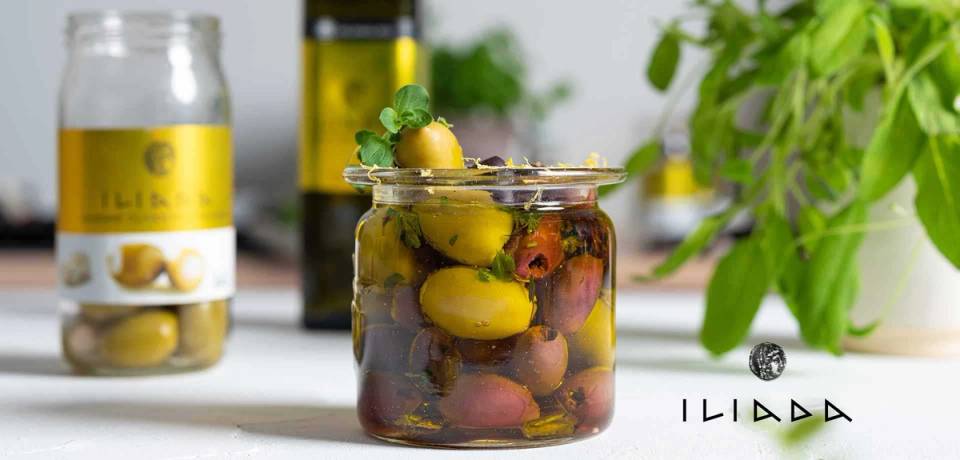 Et glass med oliven og urter, omgitt av olivenolje og grønne planter, på et kjøkkenbord.