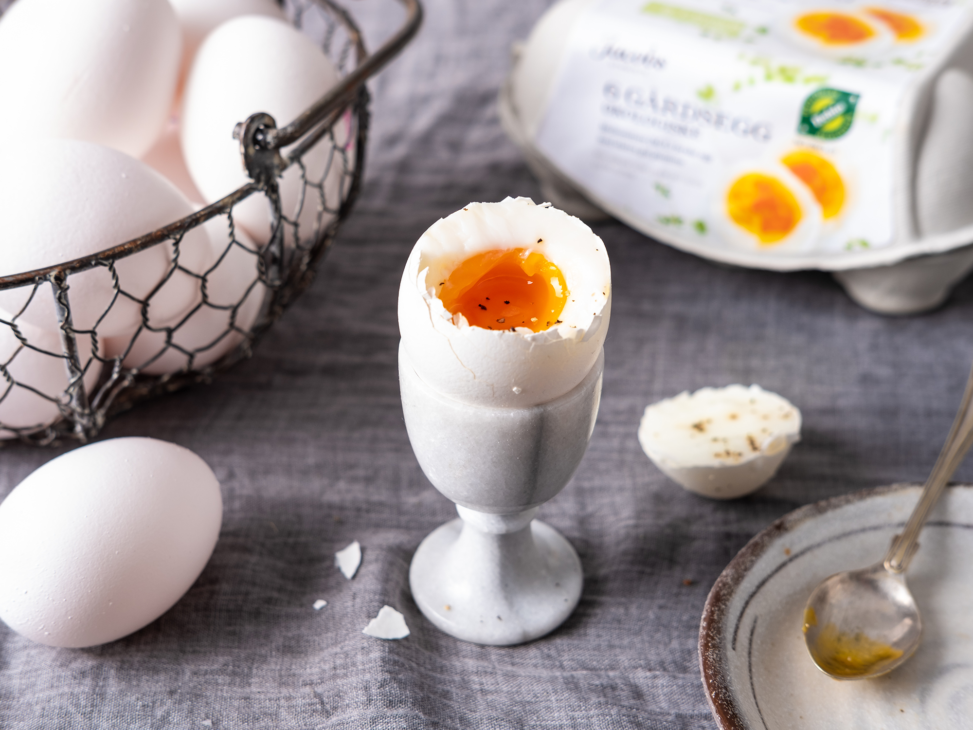 Et halvt kokt egg i et eggglass med en flytende plomme, omgitt av eggeskall og eggemballasje på en treoverflate.