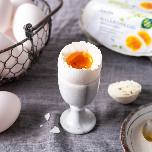 Et halvt kokt egg i et eggglass med en flytende plomme, omgitt av eggeskall og eggemballasje på en treoverflate.