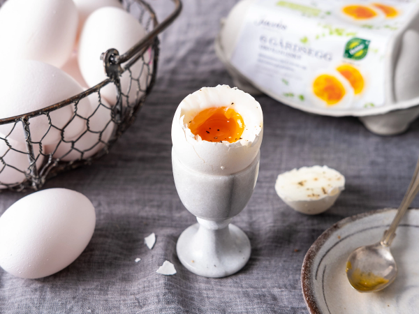 Et halvt kokt egg i et eggglass med en flytende plomme, omgitt av eggeskall og eggemballasje på en treoverflate.