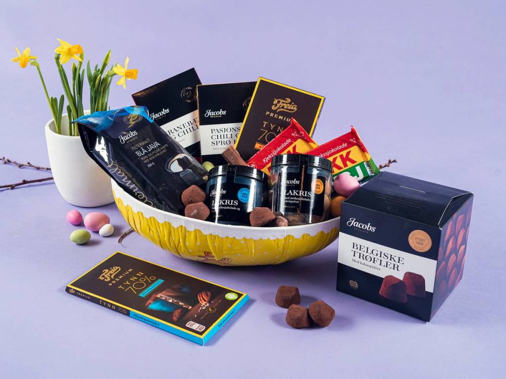 Påskeegg fylt med gourmet sjokolade og konfekt i en gul bolle, med en hvit vase med gule blomster i bakgrunnen.