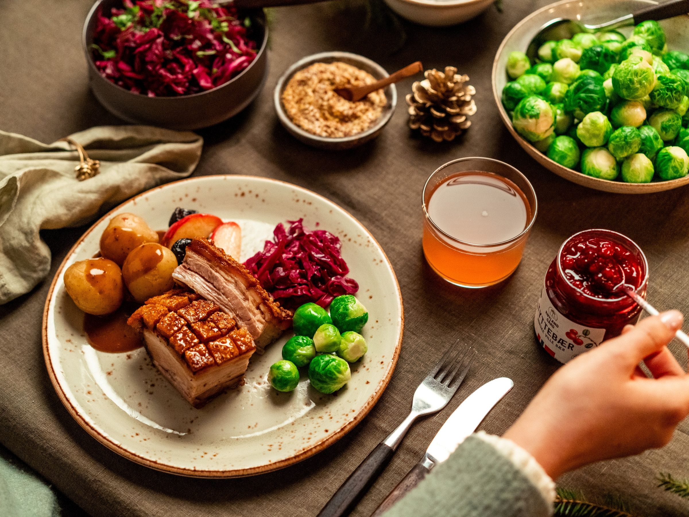 Et festmåltid med ribbe, rosenkål, julekaker, og en glass med julebrus på et bord med bestikk og pynt, sett fra oven.