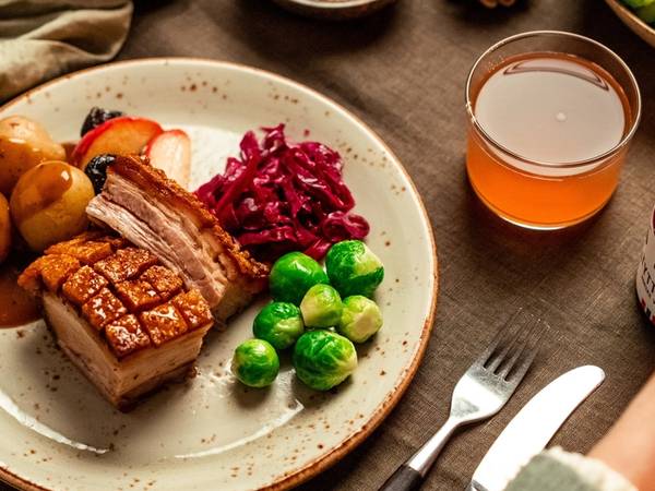 Et festmåltid med ribbe, rosenkål, julekaker, og en glass med julebrus på et bord med bestikk og pynt, sett fra oven.