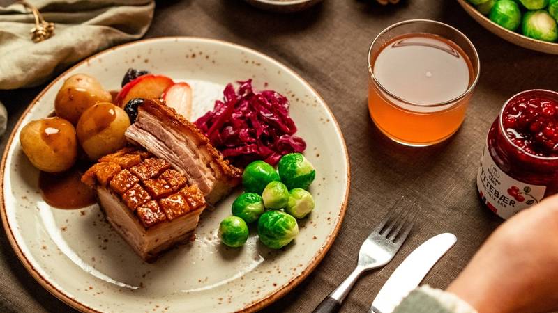 Et festmåltid med ribbe, rosenkål, julekaker, og en glass med julebrus på et bord med bestikk og pynt, sett fra oven.