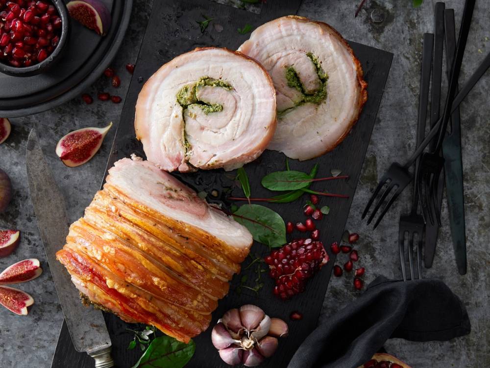 Porchetta med sprø svor og fylt med urter, skåret opp på et fat med bær og urter rundt.
