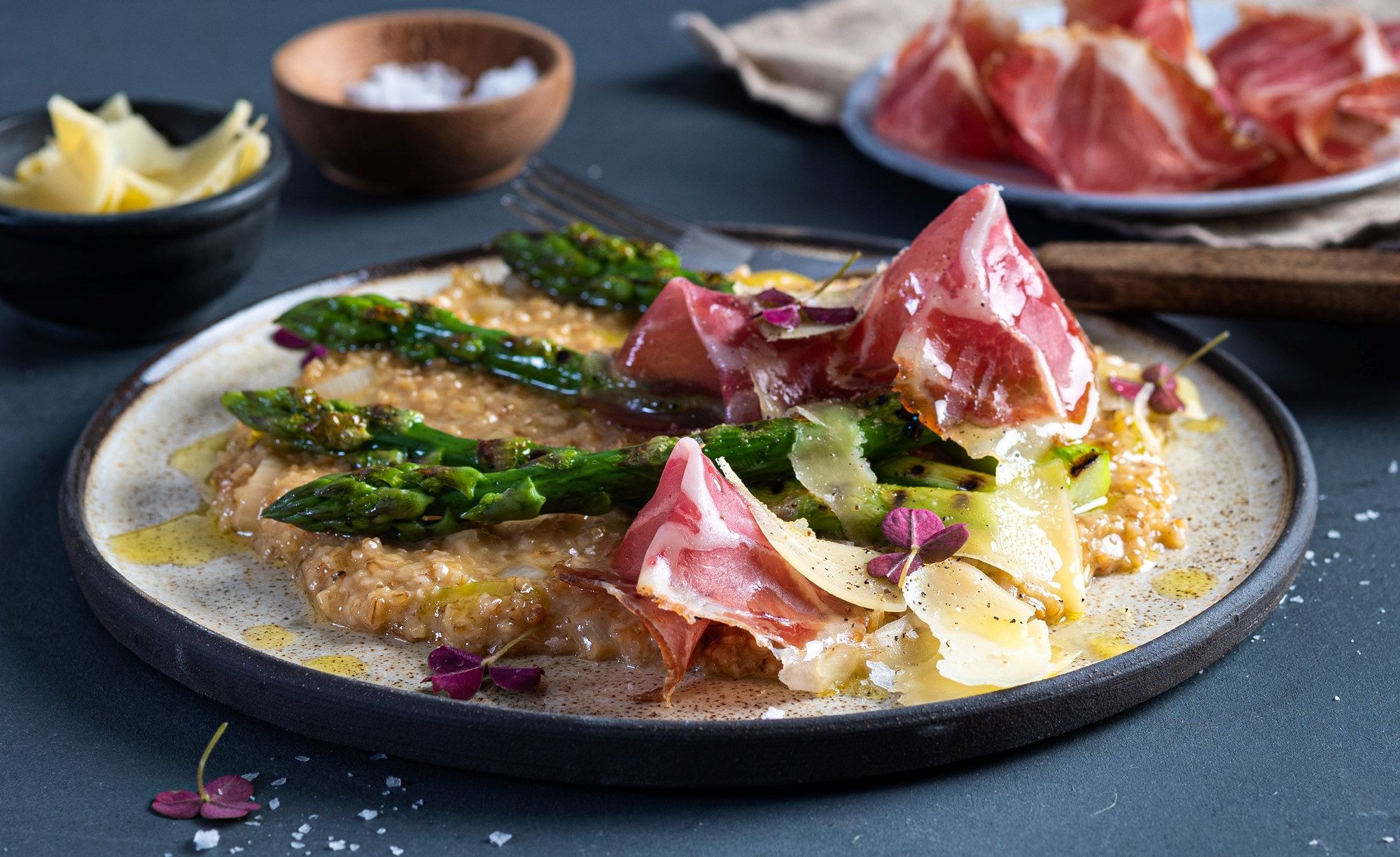 Havreotto med asparges, skinke og potetmos på en tallerken, med små boller og skinke i bakgrunnen.