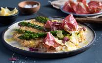 Havreotto med asparges, skinke og potetmos på en tallerken, med små boller og skinke i bakgrunnen.