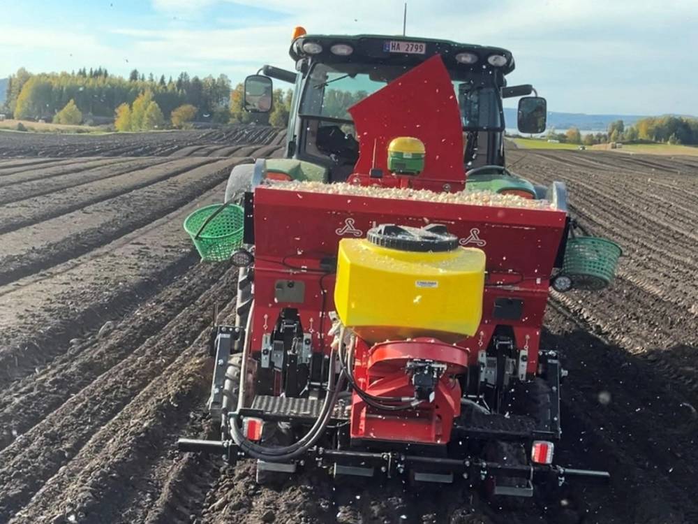 En jordbruksmaskin kjører over en åker med nyplantede rader, med åpen himmel og trær i bakgrunnen.
