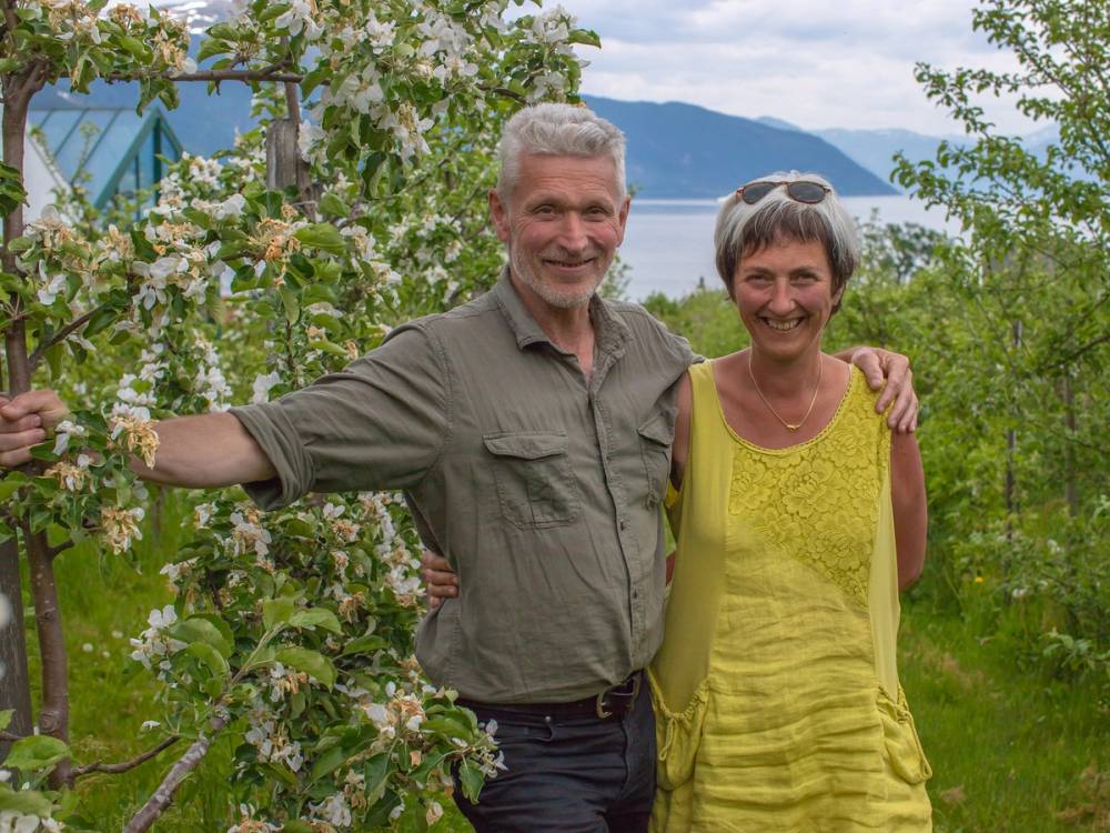 To personer står sammen i en hage med blomstrende busker og vann i bakgrunnen, en av dem har armen rundt den andre.