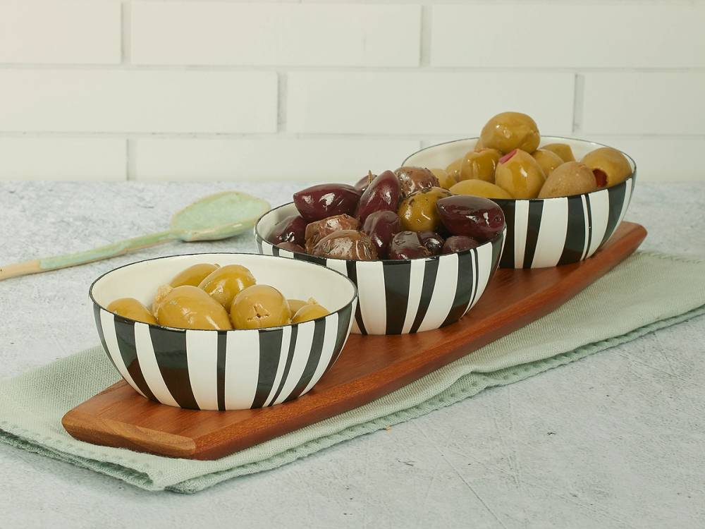 Tre skåler med ulike typer oliven på en lang treplate, plassert på et kjøkkenbord med hvit vegg i bakgrunnen.