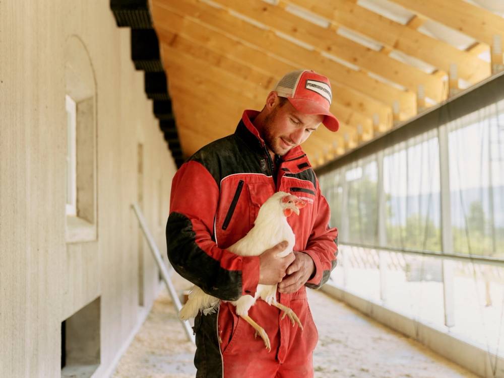 En person i rød og svart jakke holder en liten hund i armene inne i en innendørs gang med store vinduer og treverk i taket.