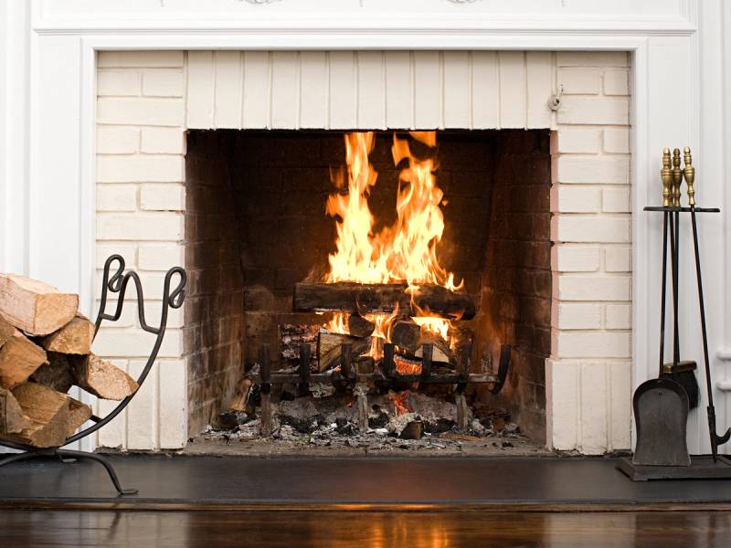 Flammende ild i en peis med vedstabler til venstre og peisspade til høyre, i et rom med hvit murvegg.