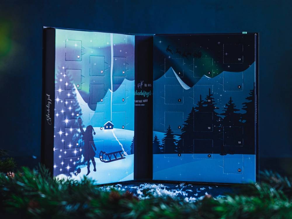 To kalendere med vinter- og juletema, med snø, juletre og en person som går med en hund, satt opp mot en mørk bakgrunn.