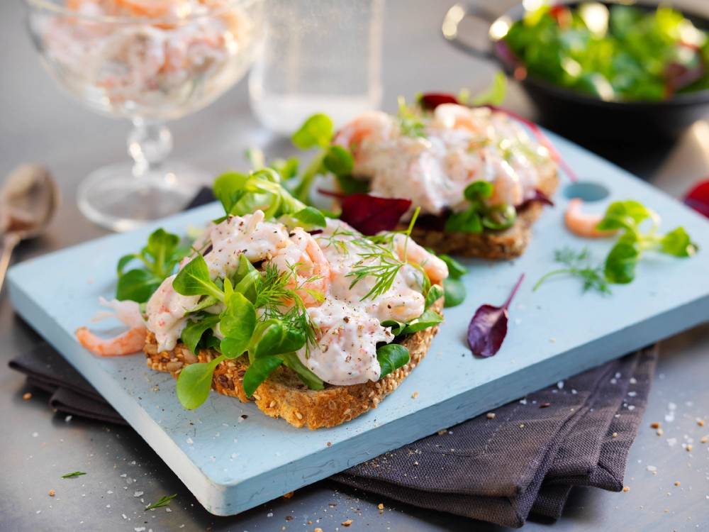 Rekesalat servert på små brødskiver med grønne urter og en kremet dressing, plassert på en lys blå serveringsplate.